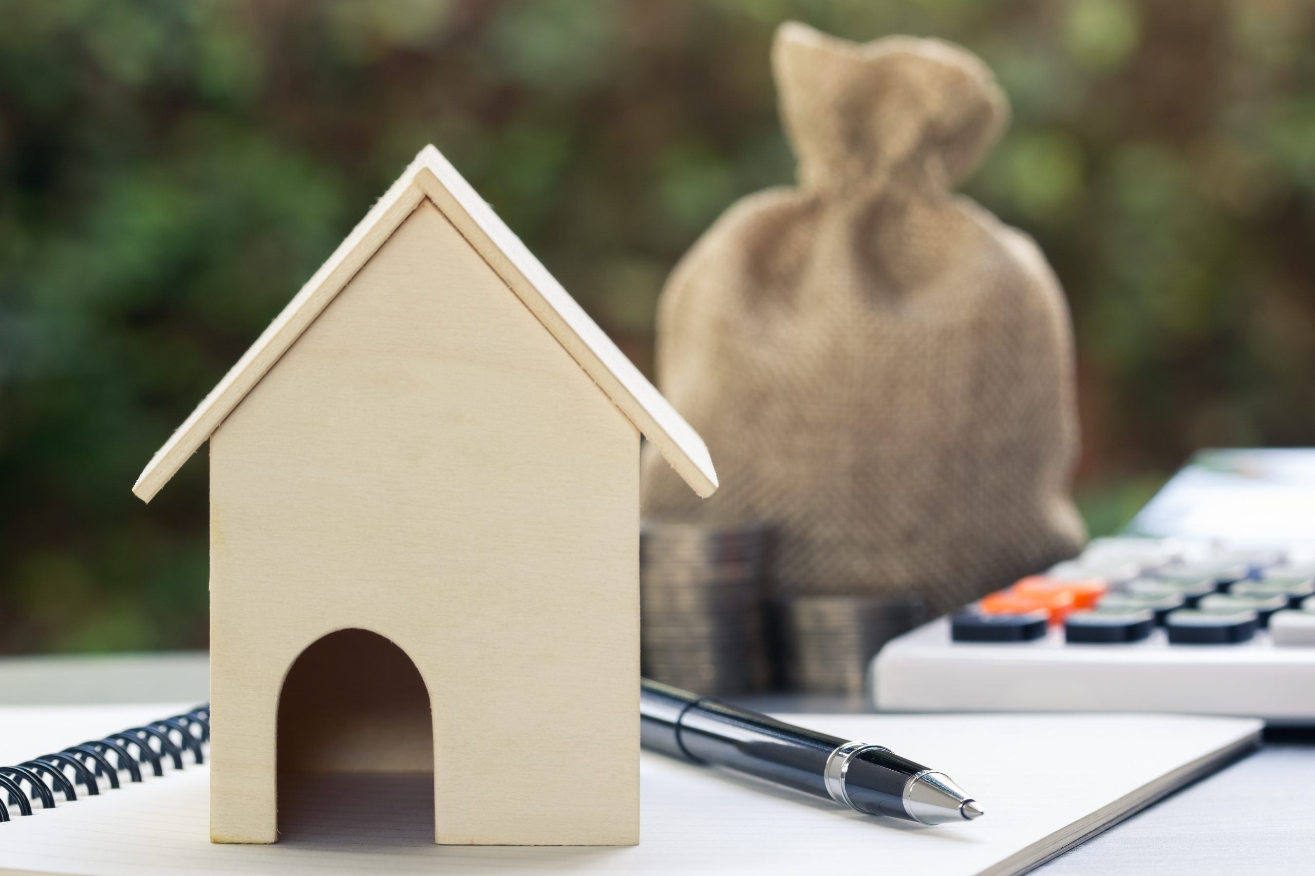 Home loans, cheap home projects, first homes to start a family concept. A small house model on notebook and pen with blurred money bag, stacked coins and calculator as background. Loan, mortgage.